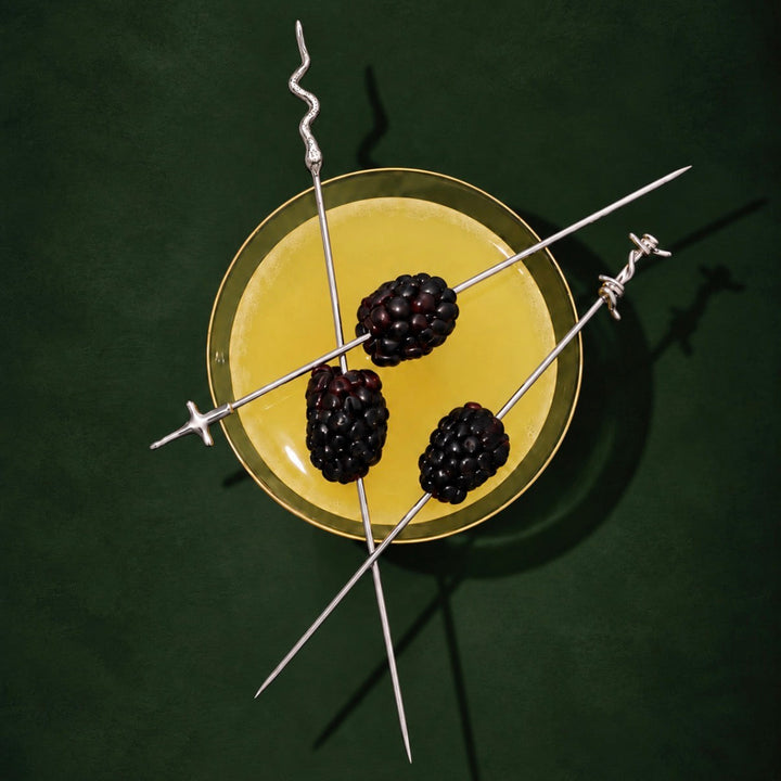 Three long silver skewers with barbed wire ends are arranged over a yellow drink in a clear glass, each threaded with a blackberry, on a dark green background.