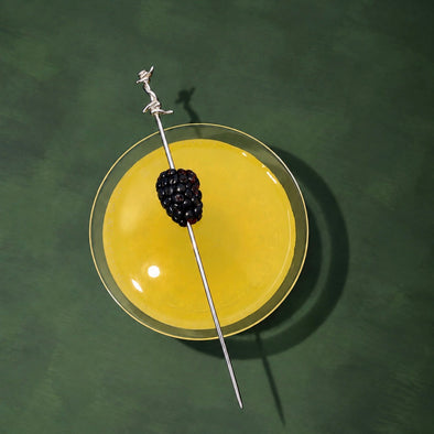A top-down view of a yellow cocktail in a clear coupe glass, garnished with a single blackberry on a silver skewer, set against a dark green textured background.