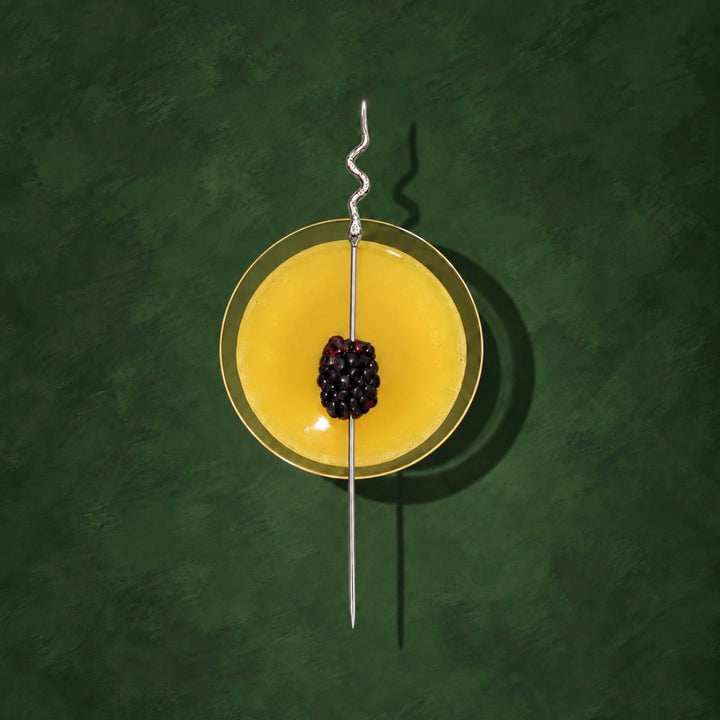 A top-down view of a yellow gelatin dessert in a round glass dish, garnished with a single blackberry on a long metal skewer, set against a dark green textured background.