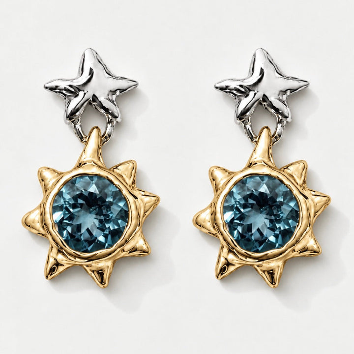 A pair of gold and silver dangle earrings featuring a central faceted blue gemstone in a spiky sunburst setting, with a textured silver star-shaped post above, shown on a light gray background.