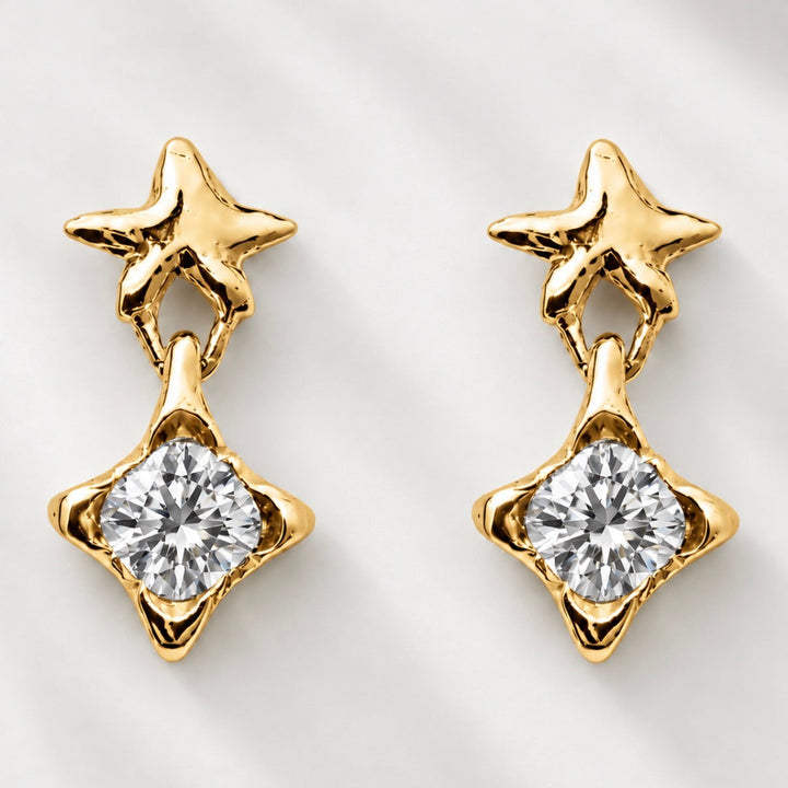 A pair of gold star-shaped dangle earrings with a clear round-cut gemstone in a four-pointed star setting, displayed on a light gray textured background.