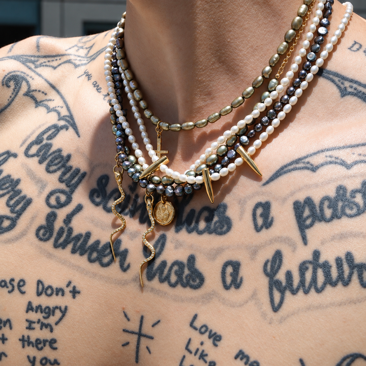 A close-up of a person's neck and upper chest wearing a layered gothic-style necklace. The necklace features white pearl beads, metallic gold beads, dark gray beads, gold spikes, a gold snake charm, and a gold coin charm.
