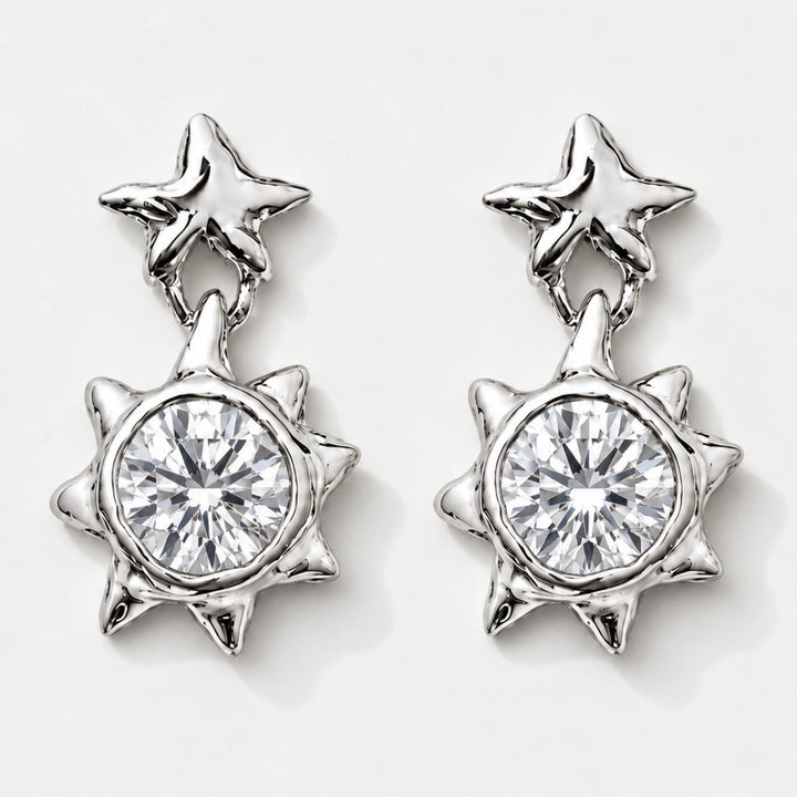 A pair of silver-toned dangle earrings featuring a star-shaped post with a dangling spiky sunburst pendant, each set with a clear round-cut gemstone, shown on a light gray background.