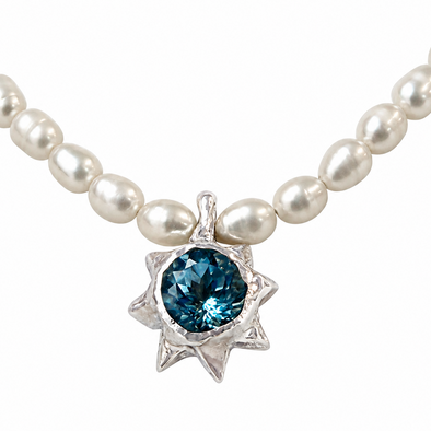A pearl necklace with a silver sunburst pendant featuring a round, faceted blue gemstone, shown against a plain white background.