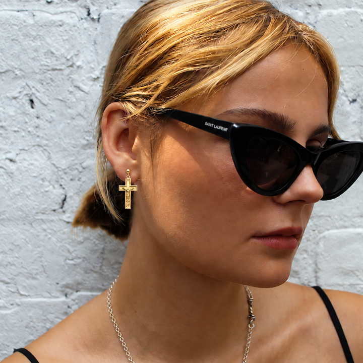 A woman with blonde hair tied back wears black cat-eye sunglasses with white 'MILANO' branding on the temple, standing against a white painted brick wall.