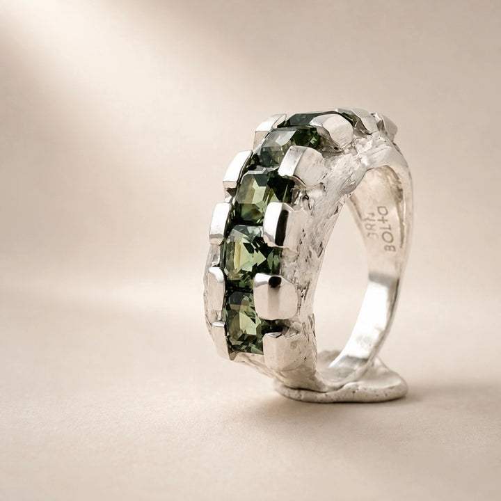 A silver ring with a textured, organic band and a row of faceted green gemstones, photographed on a warm beige background. The inside of the band is stamped with 'OTOR'.