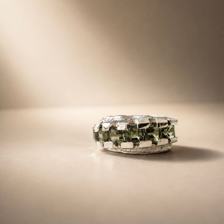 A silver ring featuring a large, faceted green gemstone, photographed on a warm beige surface with a matching background.