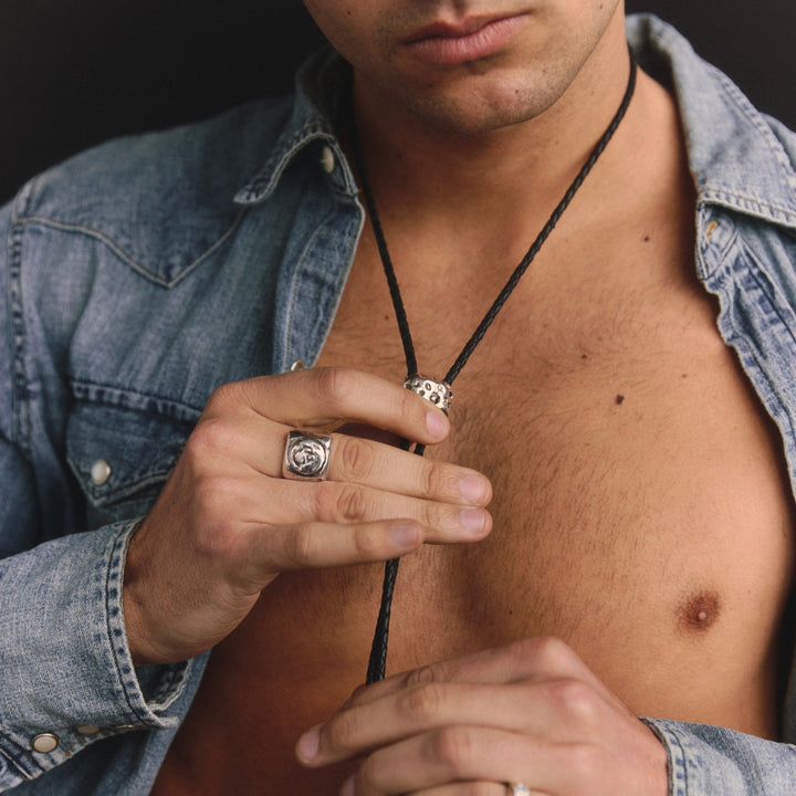 Man wearing a denim jacket with a necklace and ring, against a dark background