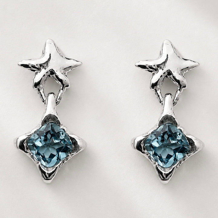 A pair of silver-toned dangle earrings featuring a small star-shaped stud at the top and a larger faceted star drop with a light blue gemstone at the bottom, set against a plain light background.