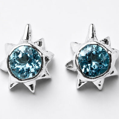 A pair of silver starburst stud earrings featuring round, faceted light blue gemstones, set against a clean white background.