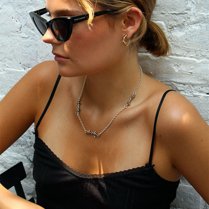 A woman with blonde-highlighted hair wearing black cat-eye sunglasses, a silver necklace, and a black spaghetti-strap top, posing against a white brick wall.