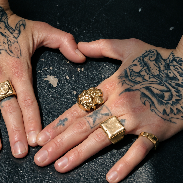 A close-up of tattooed hands wearing multiple chunky gold rings, including square bezel-set and sculptural floral designs, resting on a dark textured surface.