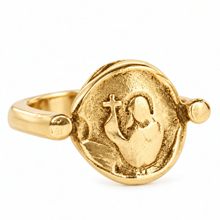 A gold-toned ring featuring a textured, circular medallion with a raised figure holding a cross, photographed on a clean white background.