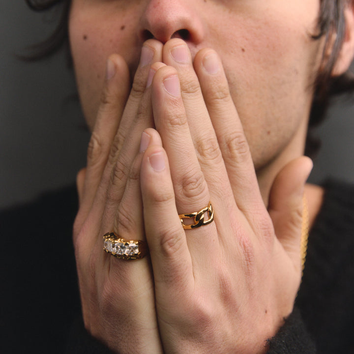 Person with two gold rings on fingers covering mouth against a dark background