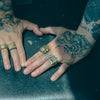Two hands with tattoos and gold rings on a dark surface