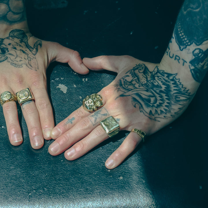 Two hands with tattoos and gold rings on a dark surface