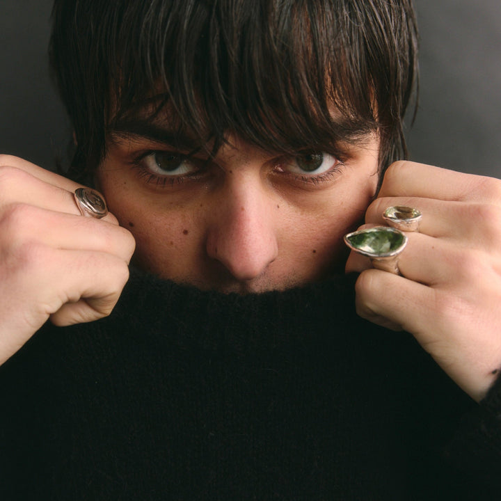 Person wearing large rings with dark hair and a black sweater against a gray background