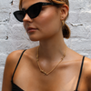 A woman with blonde hair in a low bun wearing black cat-eye sunglasses, a gold necklace, and a gold hoop earring, posing against a white brick wall.