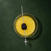 A top-down view of a yellow cocktail in a clear coupe glass, garnished with a blackberry on a metal skewer, set against a dark green background.