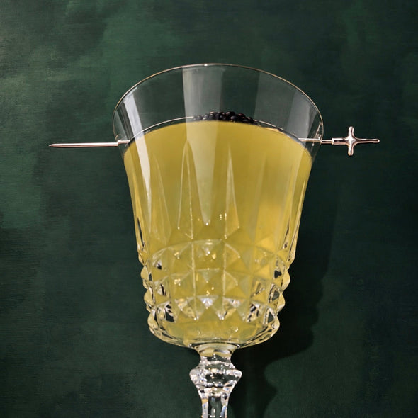 A clear cut-glass goblet with a stem, filled with a pale yellow liquid and garnished with a thin metal pick on both sides, set against a dark green textured background.