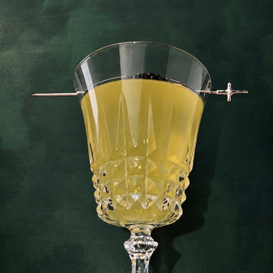 A clear cut-glass goblet with a stem, filled with a pale yellow liquid and garnished with a thin metal pick on both sides, set against a dark green textured background.