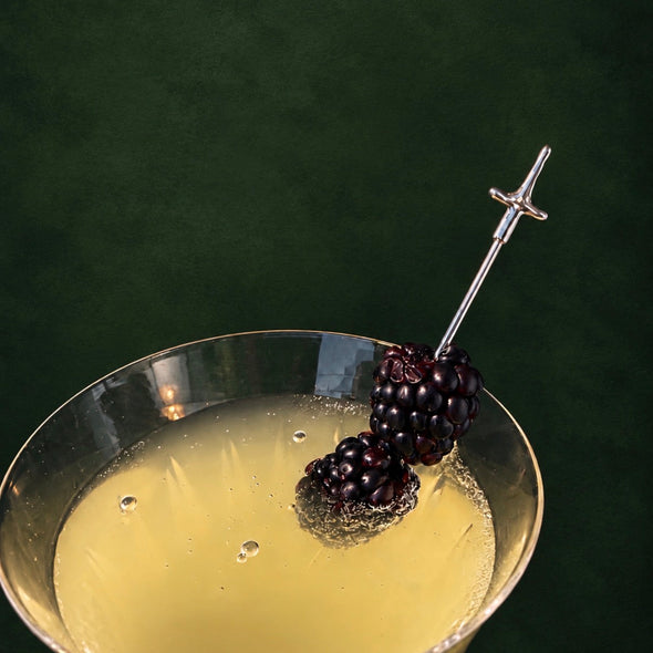 A close-up of a cocktail in a coupe glass with a silver sword-shaped garnish topped with two blackberries, set against a dark green background.