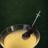 A close-up of a cocktail in a coupe glass with a silver sword-shaped garnish topped with two blackberries, set against a dark green background.