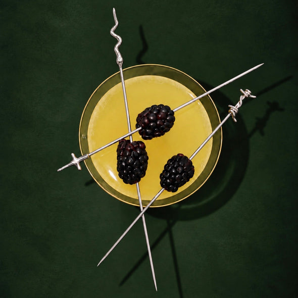 Three long silver skewers with barbed wire ends are arranged over a yellow drink in a clear glass, each threaded with a blackberry, on a dark green background.