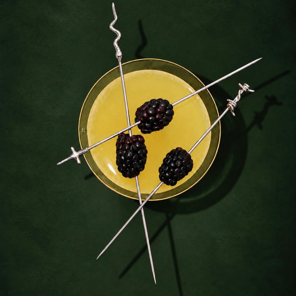 Three long silver skewers with barbed wire ends are arranged over a yellow drink in a clear glass, each threaded with a blackberry, on a dark green background.