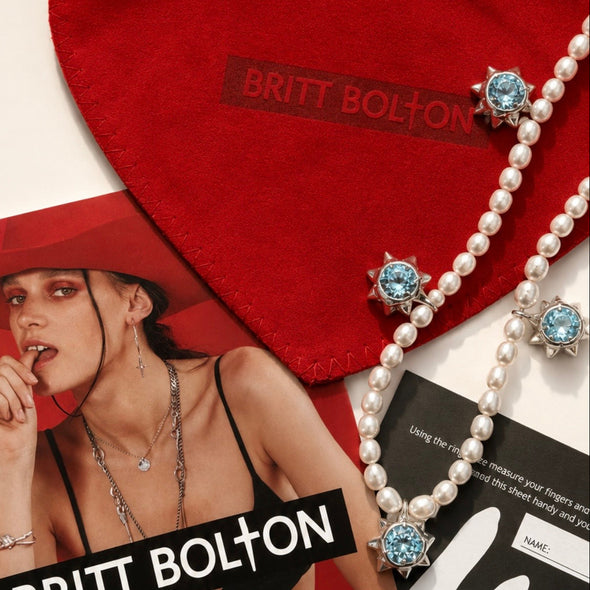 A close-up flat lay of a pearl necklace with light blue gemstone accents, shown alongside a red heart-shaped Britt Bolton pouch and printed magazine pages. The Britt Bolton brand name is visible on the pouch and in the magazine.