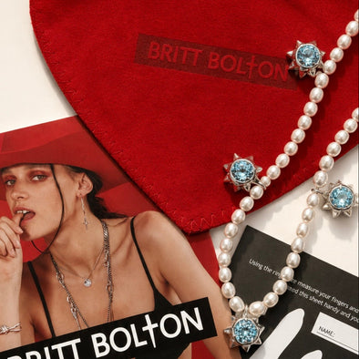 A close-up flat lay of a pearl necklace with light blue gemstone accents, shown alongside a red heart-shaped Britt Bolton pouch and printed magazine pages. The Britt Bolton brand name is visible on the pouch and in the magazine.