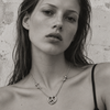 A close-up black and white portrait of a woman wearing a delicate chain necklace with a peace sign pendant and small star details, set against a textured wall.