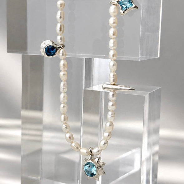 A pearl beaded necklace with silver-toned settings and blue gemstones, displayed on clear acrylic blocks against a light gray background.