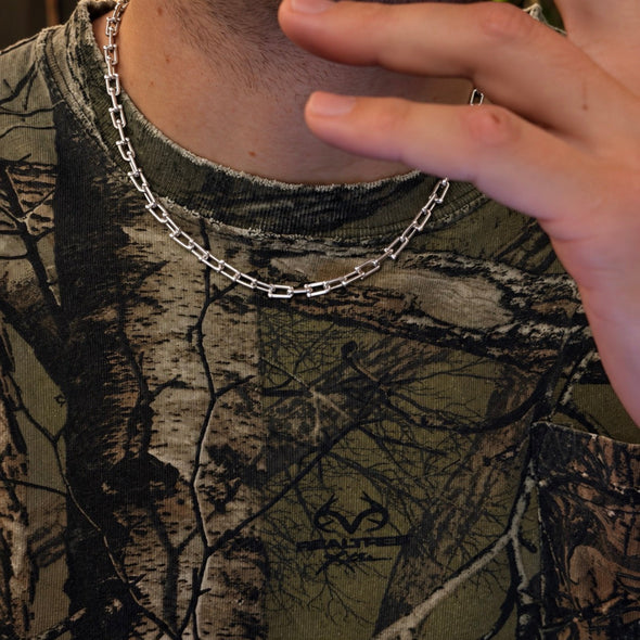 A close-up of a person wearing a silver chain necklace over a camouflage-patterned t-shirt. The person's hand is raised near their face, and a silver ring is visible on their finger.
