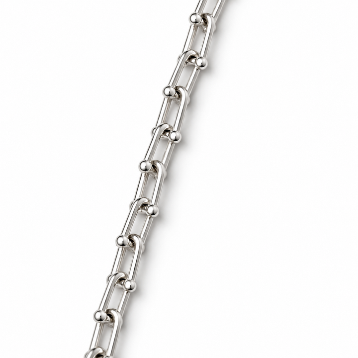 A close-up of a polished silver chain with elongated oval links and small ball accents, shown diagonally on a plain white background.