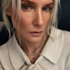 A close-up portrait of a woman with platinum blonde hair wearing a silver chain necklace with a small star charm, paired with a cream-colored knit polo shirt.