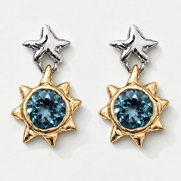 A pair of gold and silver dangle earrings featuring a central faceted blue gemstone in a spiky sunburst setting, with a textured silver star-shaped post above, shown on a light gray background.