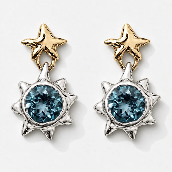 A pair of celestial dangle earrings featuring gold star-shaped studs and silver sunburst pendants with round blue gemstones, set against a clean white background.