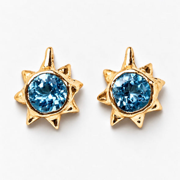 A pair of gold starburst stud earrings with round light blue gemstones, displayed on a clean white background.