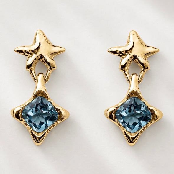 A pair of gold dangle earrings featuring a textured star-shaped post and a dangling blue faceted gemstone set in a starburst design, shown on a light gray background.