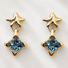 A pair of gold dangle earrings featuring a textured star-shaped post and a dangling blue faceted gemstone set in a starburst design, shown on a light gray background.