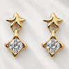 A pair of gold star-shaped dangle earrings with a clear round-cut gemstone in a four-pointed star setting, displayed on a light gray textured background.