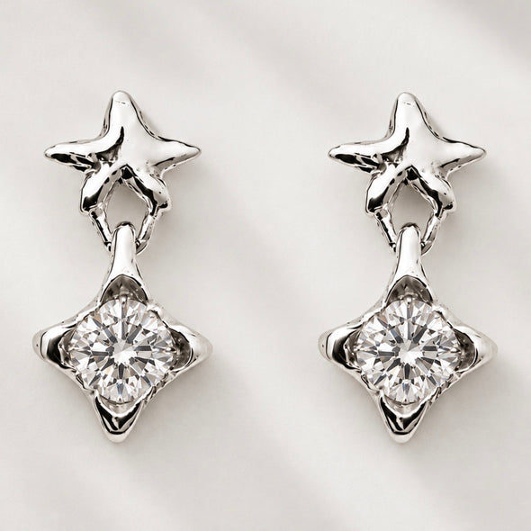 A pair of silver-toned dangle earrings featuring a small star-shaped stud at the top and a larger clear faceted gemstone in a four-pointed star setting below, displayed on a light textured background.