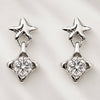 A pair of silver-toned dangle earrings featuring a small star-shaped stud at the top and a larger clear faceted gemstone in a four-pointed star setting below, displayed on a light textured background.