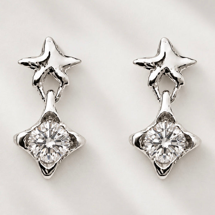 A pair of silver-toned dangle earrings featuring a small star-shaped stud at the top and a larger clear faceted gemstone in a four-pointed star setting below, displayed on a light textured background.