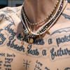 A close-up of a person's neck and upper chest wearing a layered gothic-style necklace. The necklace features white pearl beads, metallic gold beads, dark gray beads, gold spikes, a gold snake charm, and a gold coin charm.