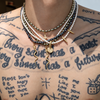 A close-up of a person's tattooed chest wearing multiple layered necklaces, including white pearl strands, black beads, and gold-toned chains with pendants.