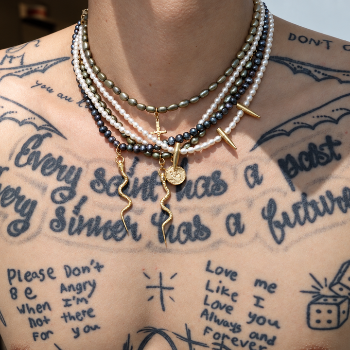 A close-up of a person's tattooed chest wearing multiple layered necklaces, including white pearl strands, black beads, and gold-toned chains with pendants.
