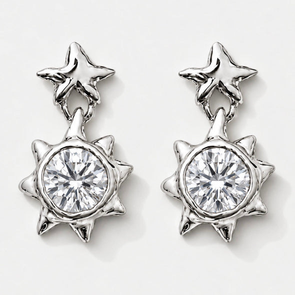 A pair of silver-toned dangle earrings featuring a star-shaped post with a dangling spiky sunburst pendant, each set with a clear round-cut gemstone, shown on a light gray background.