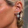 A close-up of a model's ear wearing a silver starburst earring with clear rhinestones and a small dangling olive green fabric charm, along with smaller stud earrings.